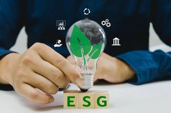 Eine Person hält eine Glühbirne mit grünen Blattsymbolen über Holzblöcken mit der Aufschrift ESG, die innovative Wege zur Verbesserung Ihres ESG-Scores symbolisieren.