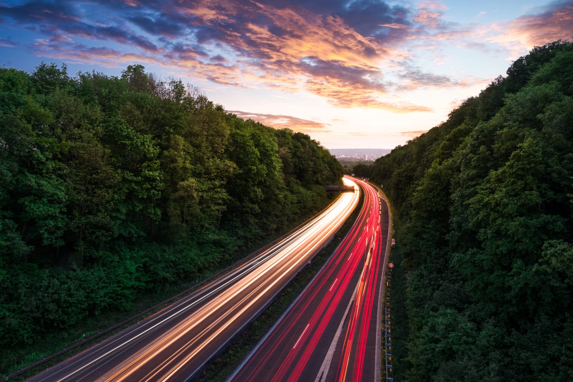Ein Foto mit Langzeitbelichtung von einer Autobahn durch einen Wald bei Sonnenuntergang, mit Lichtspuren von Autos - eine Aufnahme der Reise, die dem Verständnis von Änderungen der Reisekostenabrechnung oder der Planung für die Reisekosten 2025 sehr nahe kommt.