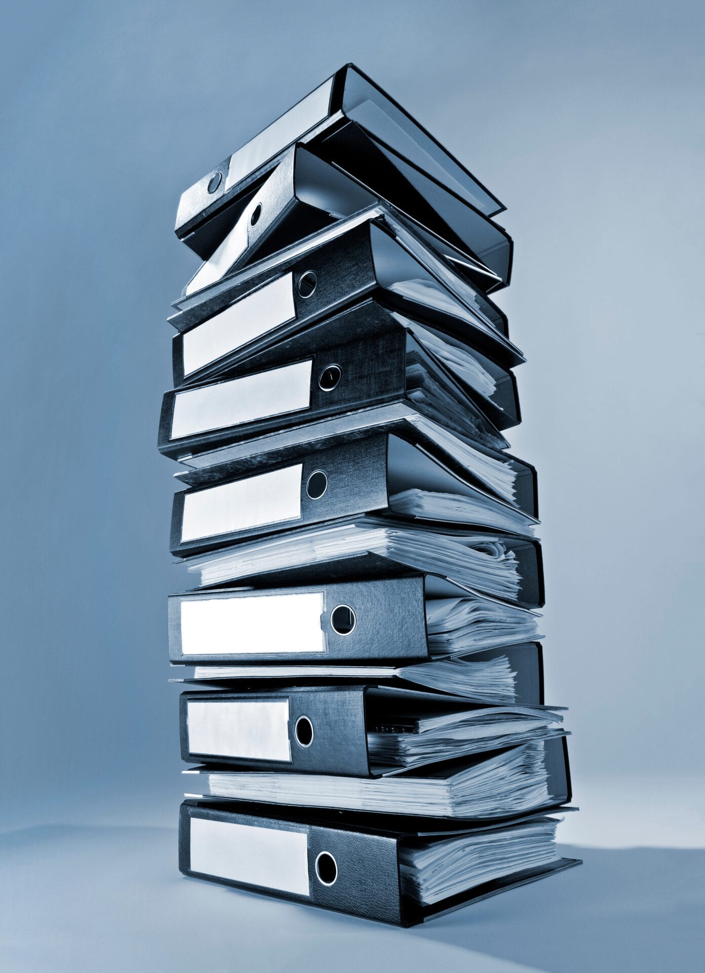 Ein hoher Stapel überladener Ordner und Mappen, gefüllt mit Papieren vor blauem Hintergrund, veranschaulicht die Aufbewahrungspflichten, die Unternehmen für eine ordnungsgemäße Dokumentenablage befolgen müssen.