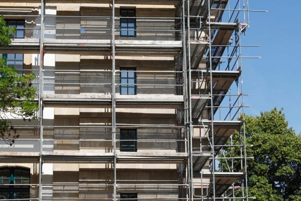 Ein Baugerüst bedeckt die Seite eines im Bau befindlichen Gebäudes und hebt energieeffizientes Bauen neben Bäumen und einem klaren blauen Himmel hervor.