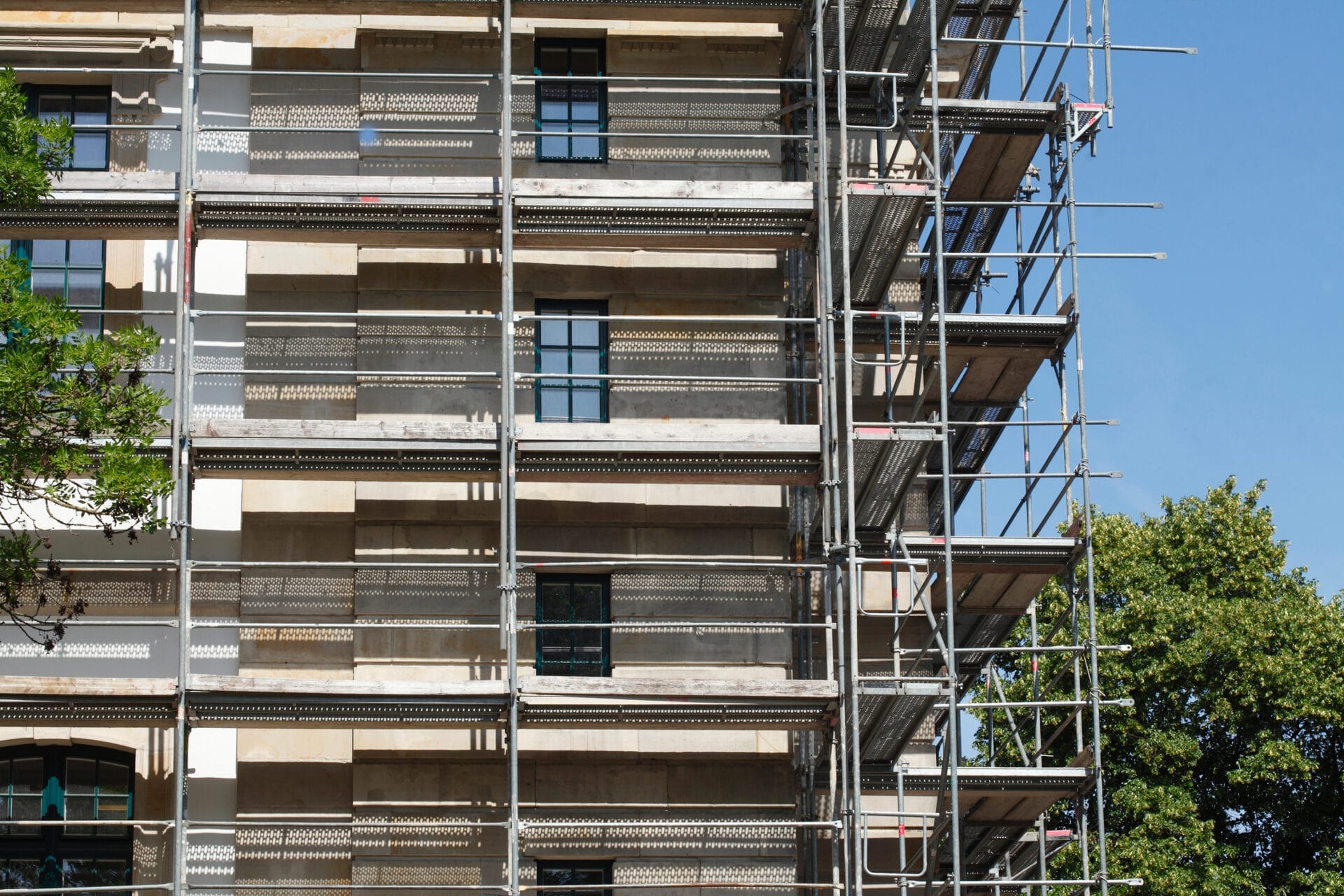 Ein Baugerüst bedeckt die Seite eines im Bau befindlichen Gebäudes und hebt energieeffizientes Bauen neben Bäumen und einem klaren blauen Himmel hervor.
